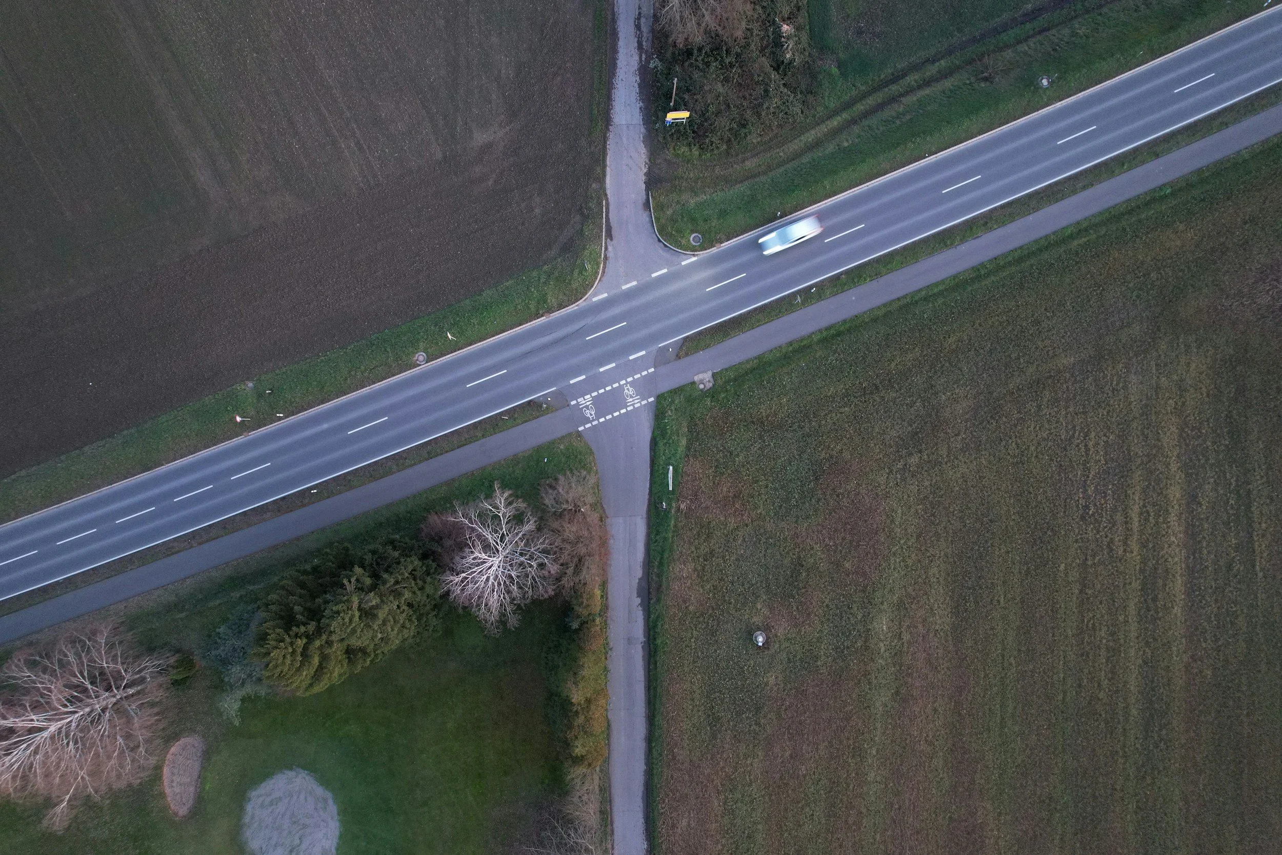 Aerial view of a rural road intersection with a car, adjacent to fields and trees.