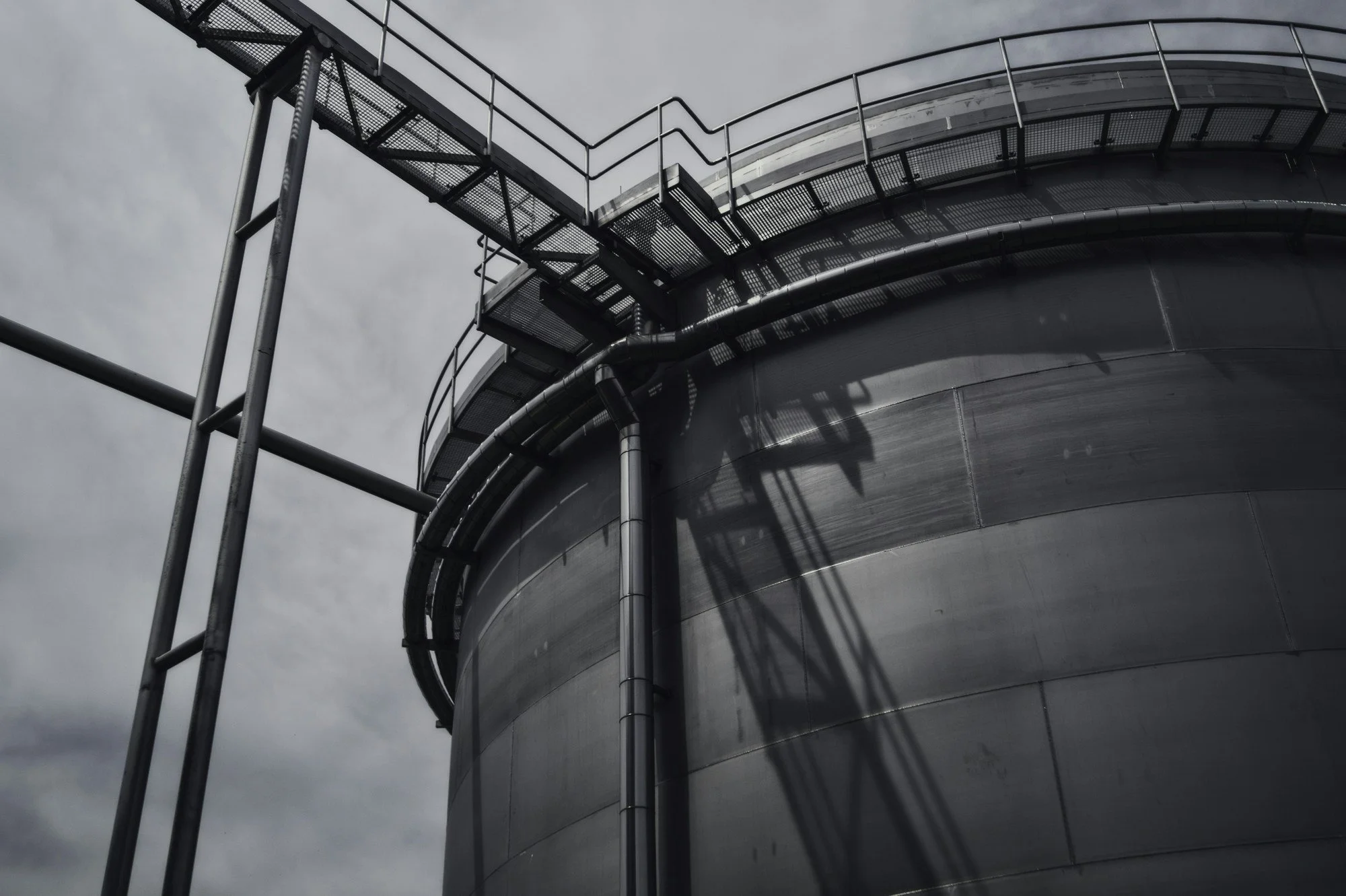 Close-up of a metallic industrial tank with pipes and metal stairs attached, under a cloudy sky.