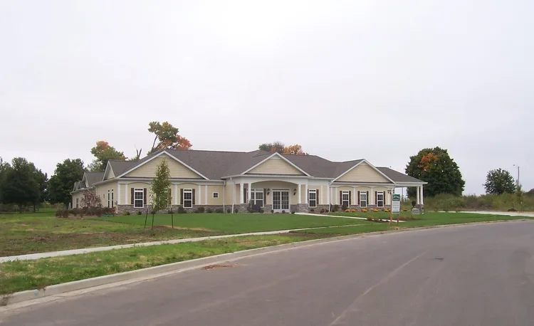 Large, light-colored house with a porch, situated on a grassy lawn near a paved road, under an overcast sky.