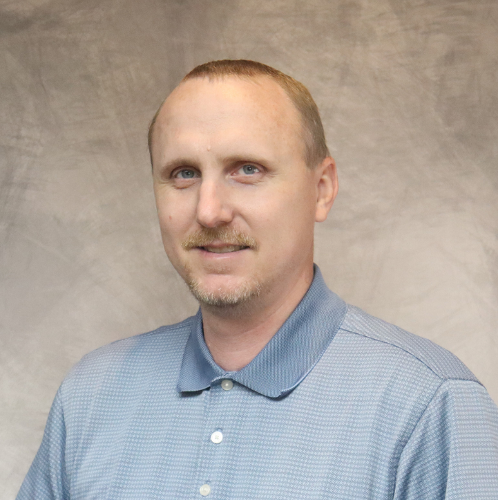 A man with short light brown hair, blue eyes, and a light beard, wearing a blue collared shirt, standing in front of a neutral background.