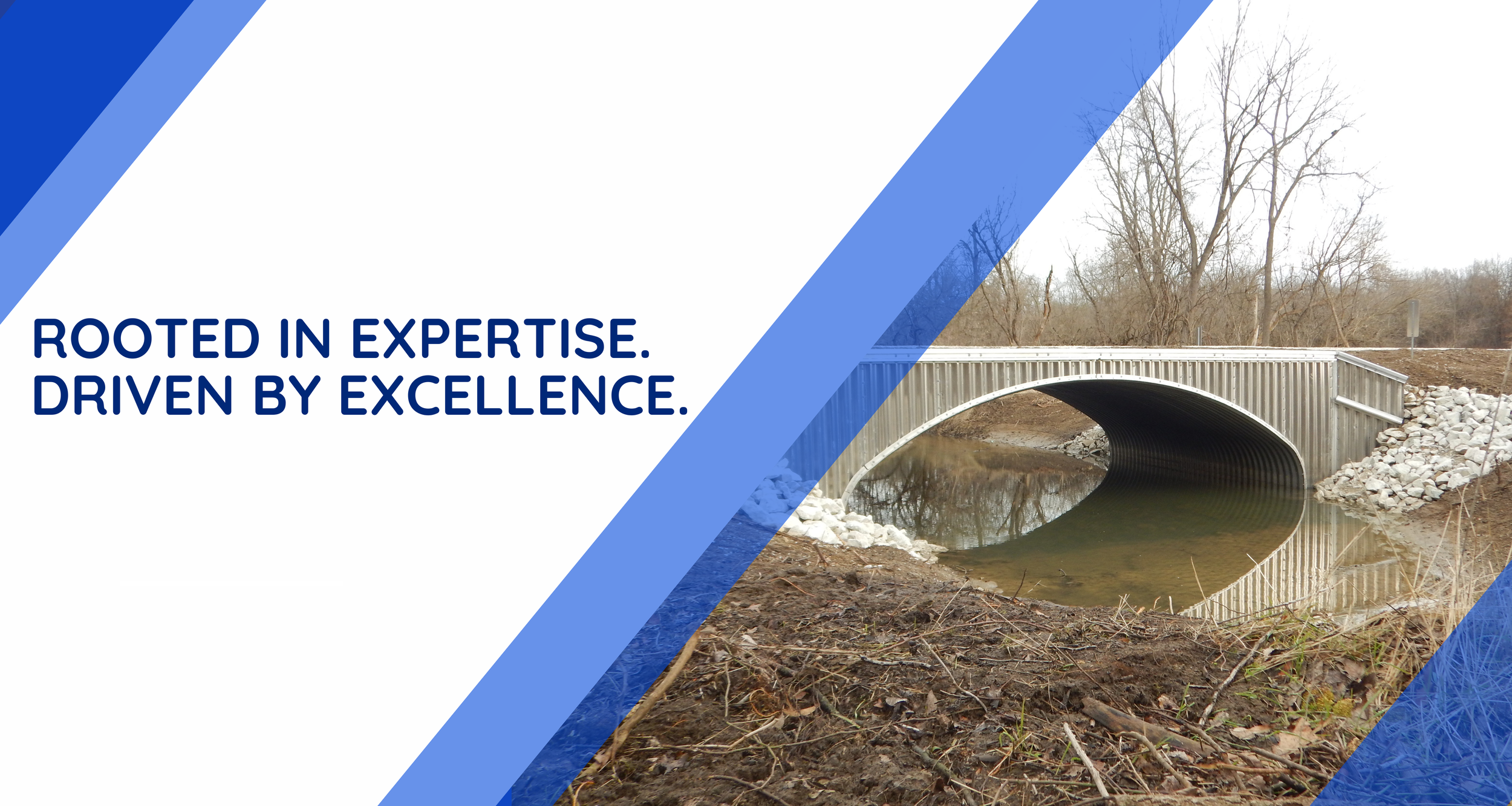 A construction photo of a small metal bridge crossing over a narrow river with a dirt bank and leafless trees in the background.