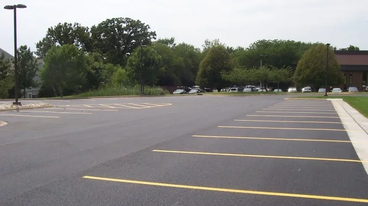 Empty parking lot with yellow painted parking spaces, surrounded by trees and a building in the background.