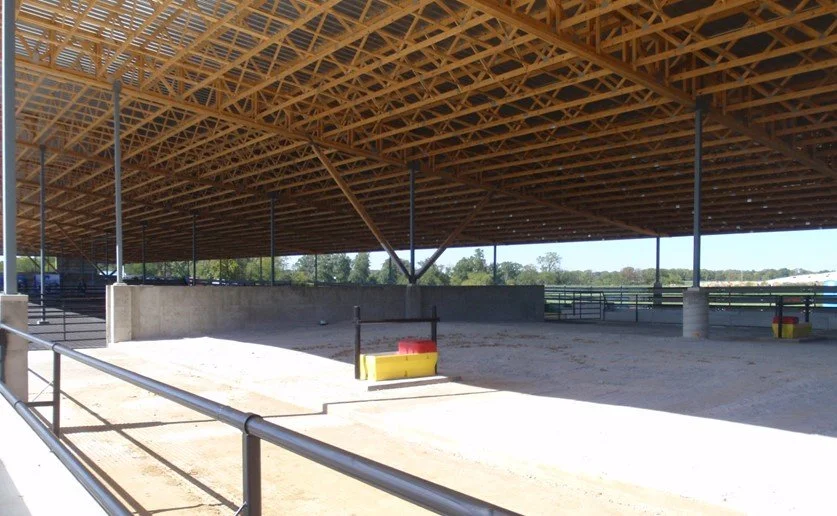 An empty covered outdoor arena with a dirt surface, metal railings, and a wooden roof structure.
