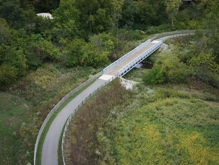 An aerial view of a curved outdoor pedestrian bridge with a ramp and railing, crossing over a small stream surrounded by green trees and grassy areas.