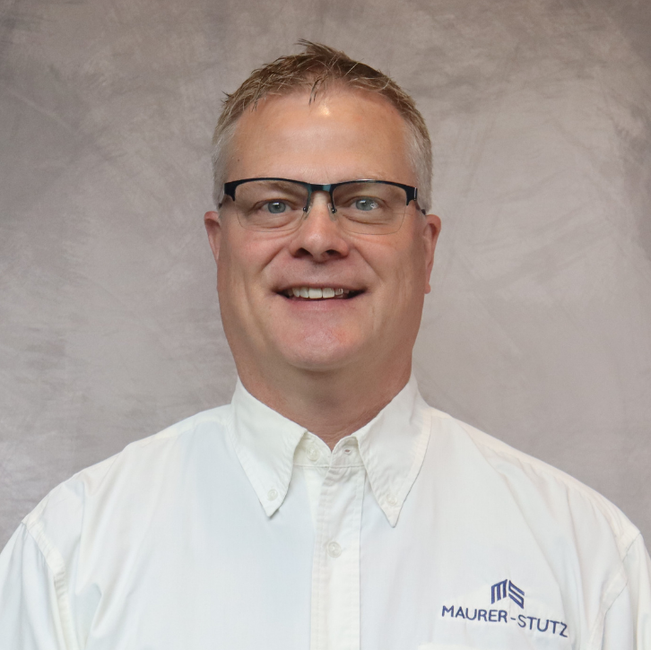 A man with glasses and short blonde hair smiling, wearing a white collared shirt with a logo that says Maurer-Stutz.