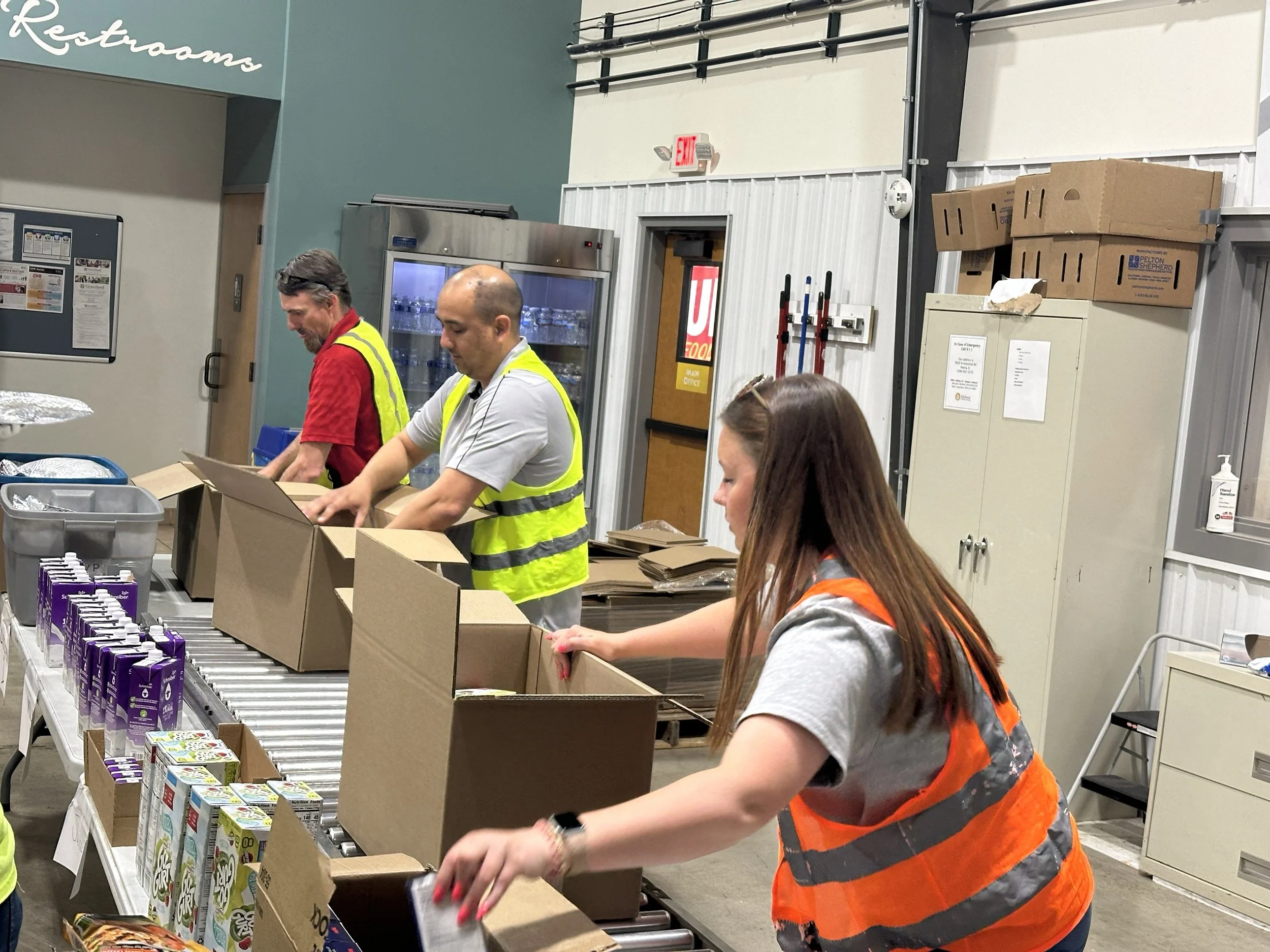 Three workers, two men and one woman, are packing boxes on a conveyor belt in a warehouse. The woman is wearing an orange safety vest, and the men are wearing yellow safety vests. There are additional boxes and packaging materials on the table.