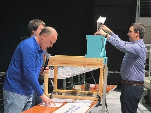 Three men at an indoor event, with one man holding a microphone and looking at a display board, another man pointing at the display, and the third man holding a stack of papers.