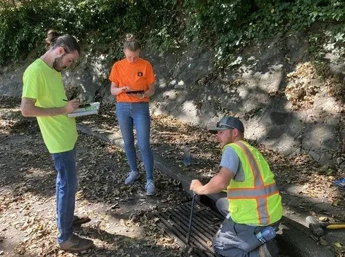 Three people working outdoors near a storm drain. 