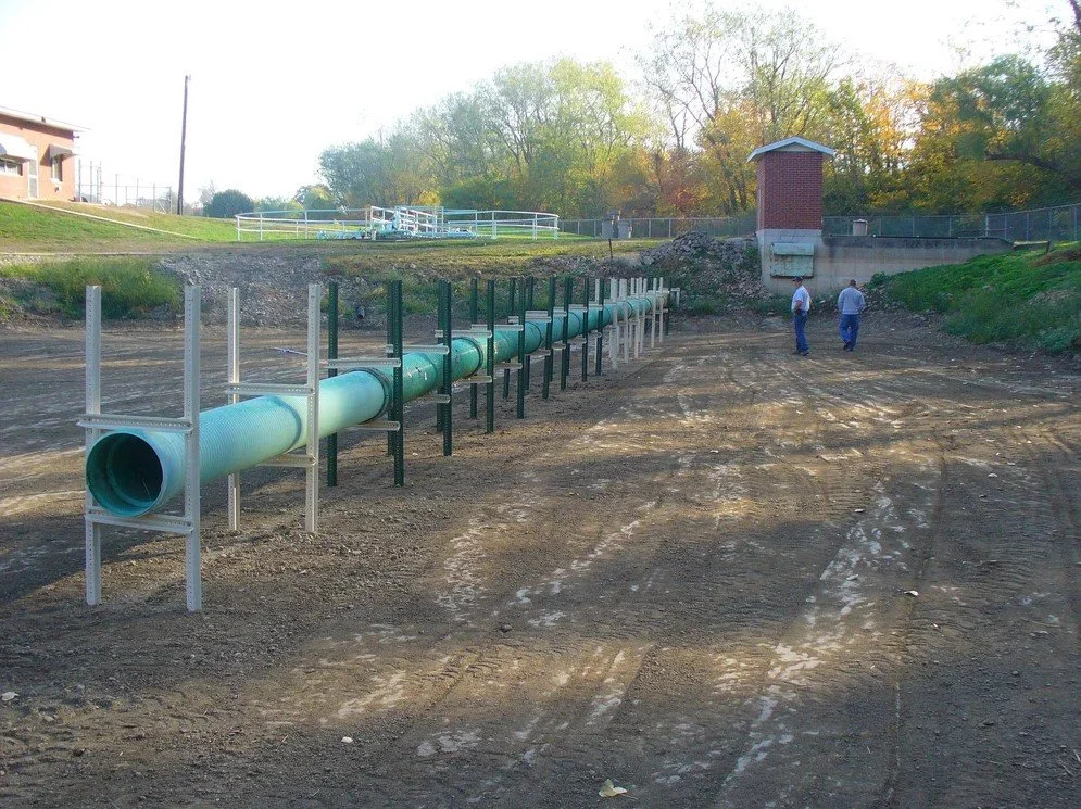 Construction site with large green pipe, two men walking, cleared dirt area, trees, and a small building in the background.