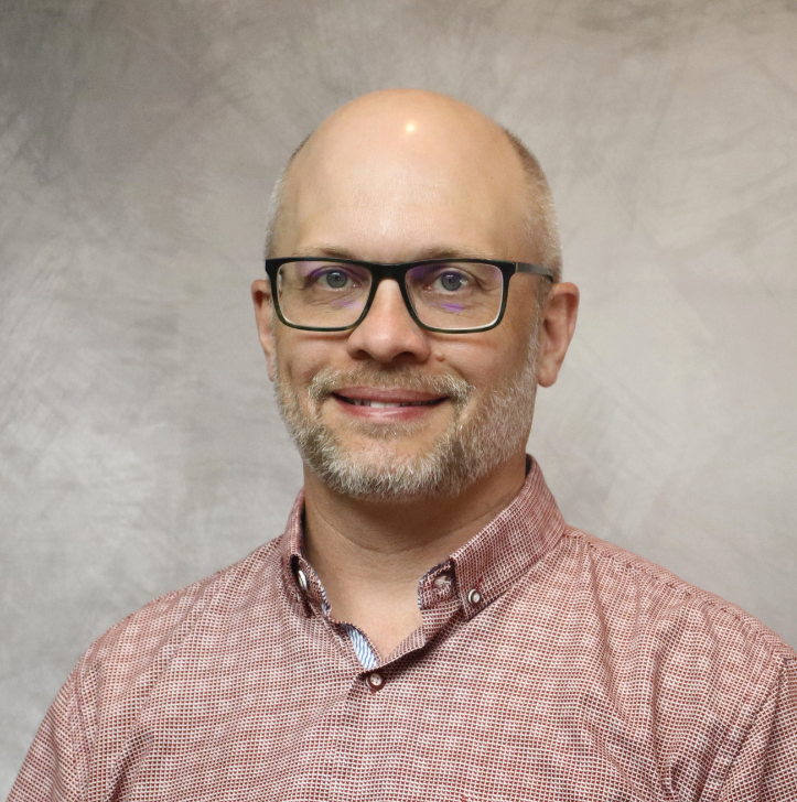 Portrait of a man with glasses, a beard, and a mustache, wearing a red checkered shirt, smiling at the camera with a neutral textured background.