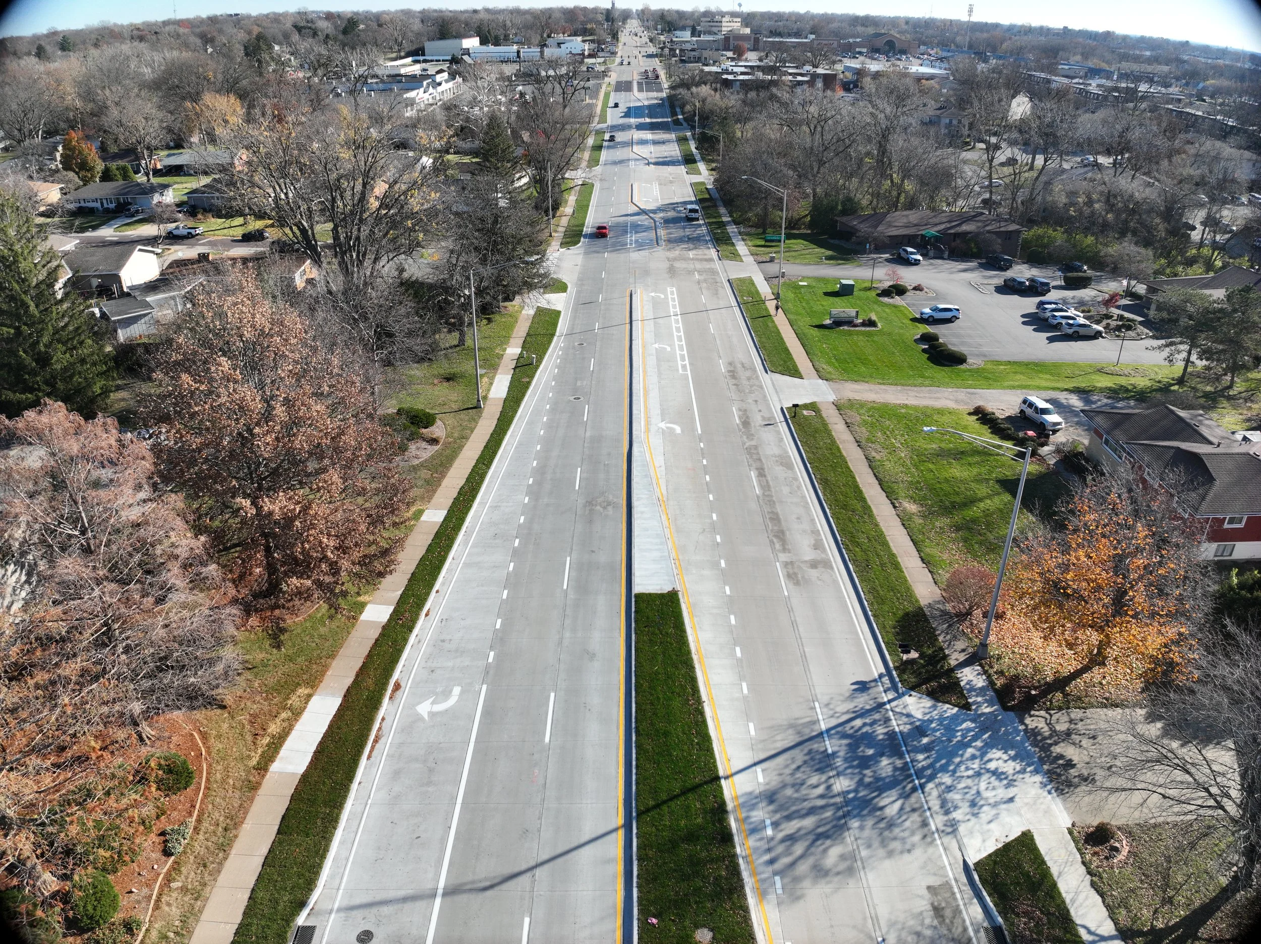 A road construction scene in a suburban neighborhood with a steamroller, traffic cones, and a 'No Parking' sign, and green trees and houses in the background.
