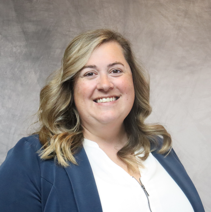A woman with wavy blonde hair, wearing a white top and navy blazer, smiling at the camera against a neutral background.