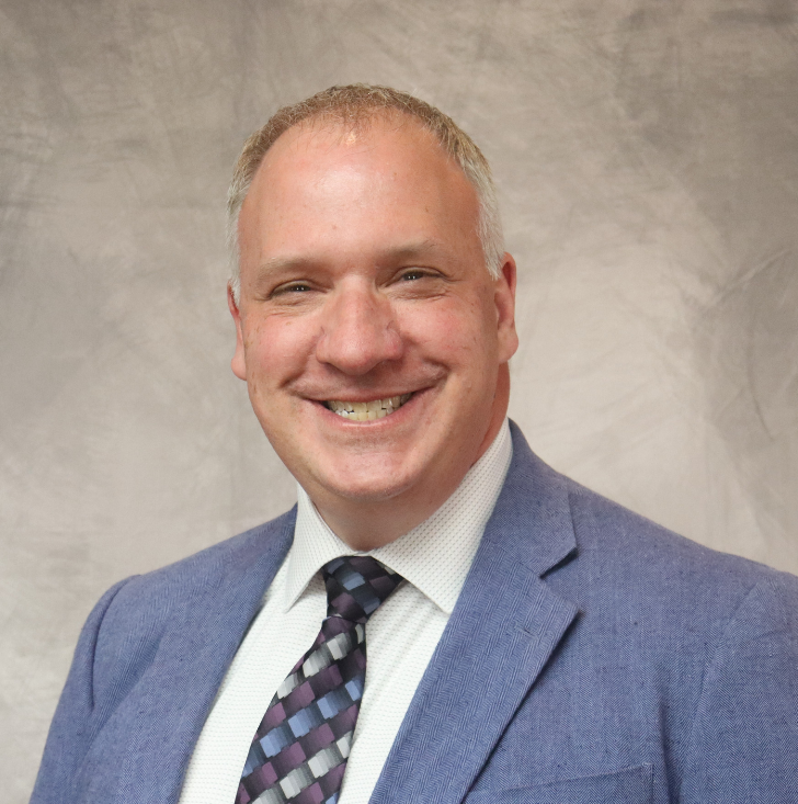 Portrait of a smiling man in a blue suit, white shirt, and patterned tie, standing against a plain background.