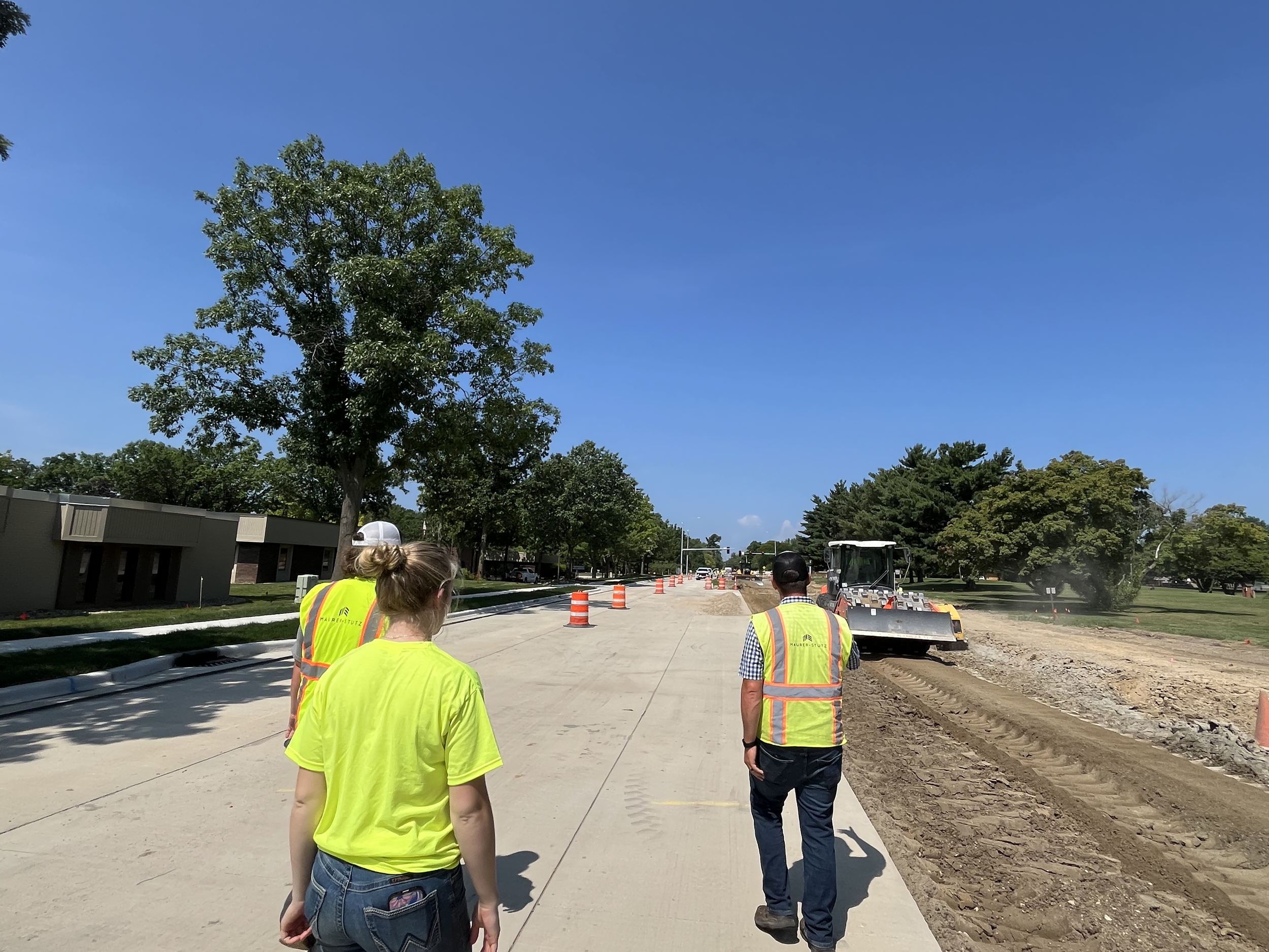Three construction workers walking on a sidewalk under construction, with a street roller and traffic cones in the background, trees lining the street, and a clear blue sky.