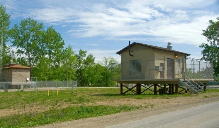 Small beige utility building raised on stilts with stairs, surrounded by trees and a dirt road.