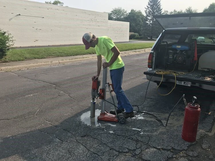 Worker in neon yellow shirt and baseball cap using specialized equipment to repair a pothole in asphalt near a parked utility vehicle.