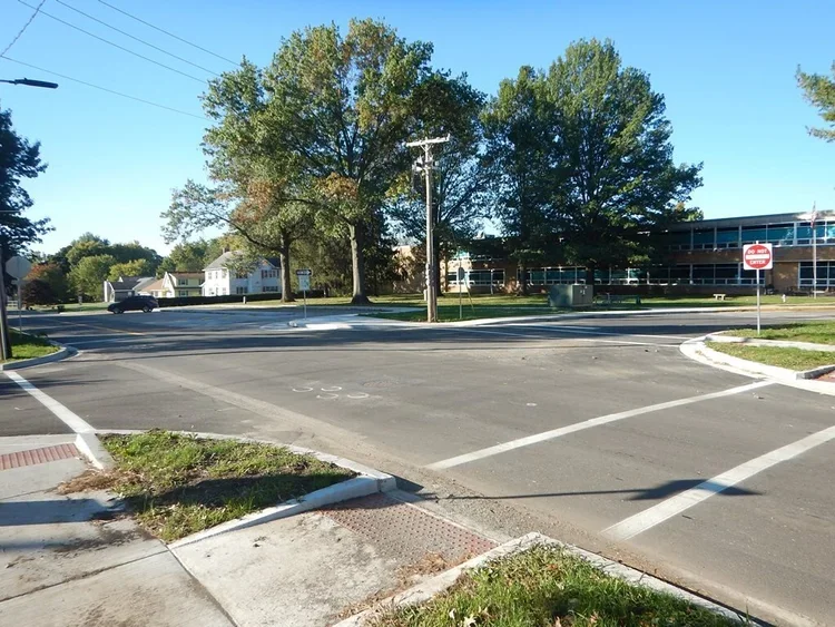 An empty four-way intersection with crosswalks, stop signs, and traffic signals, surrounded by trees, buildings, and residential houses, under a clear blue sky.