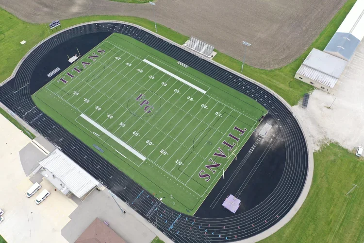 An aerial view of a football field with the word 'TITANS' painted at both ends, surrounded by a running track, and adjacent buildings and fields.
