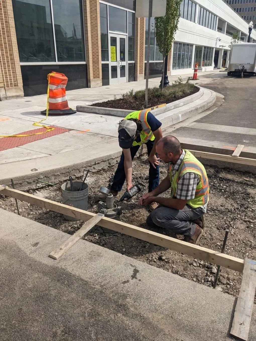 Two construction workers in safety vests working on installing or repairing a sidewalk or curb on a city street. One worker is kneeling while the other is bent over, handling tools. Construction cones and equipment are visible nearby.