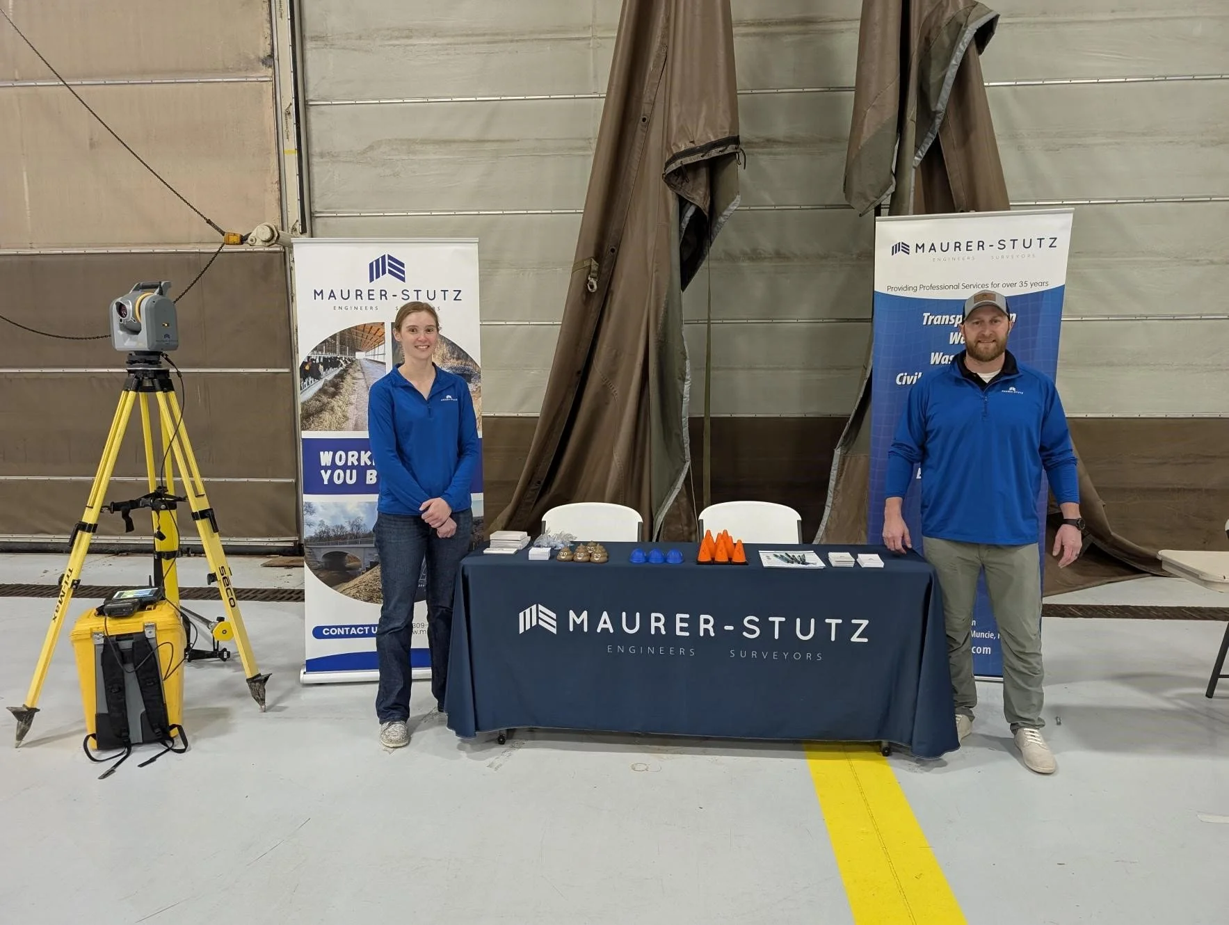 Two people standing behind a table with pamphlets and promotional items at a trade show booth for Maurer-Stutz, an engineering and surveying company. The booth is held in a large indoor space with metal walls and a yellow calibration tripod on the le