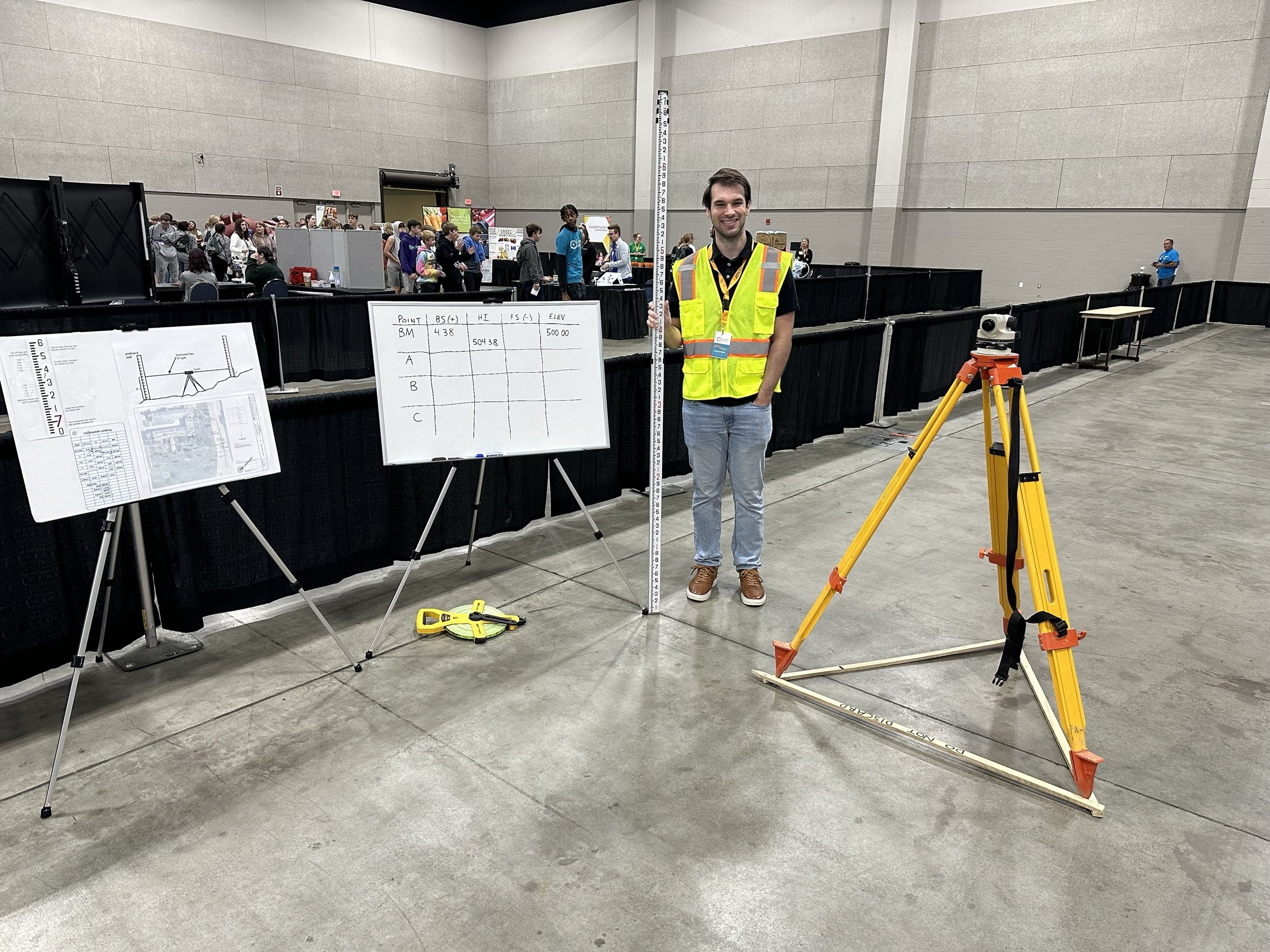 A man in a yellow safety vest standing next to a survey measuring rod and tripod at an indoor event with several booths and people in the background.