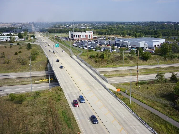 An aerial view of a highway with a few cars, an overpass, and commercial buildings in the background under a partly cloudy sky.