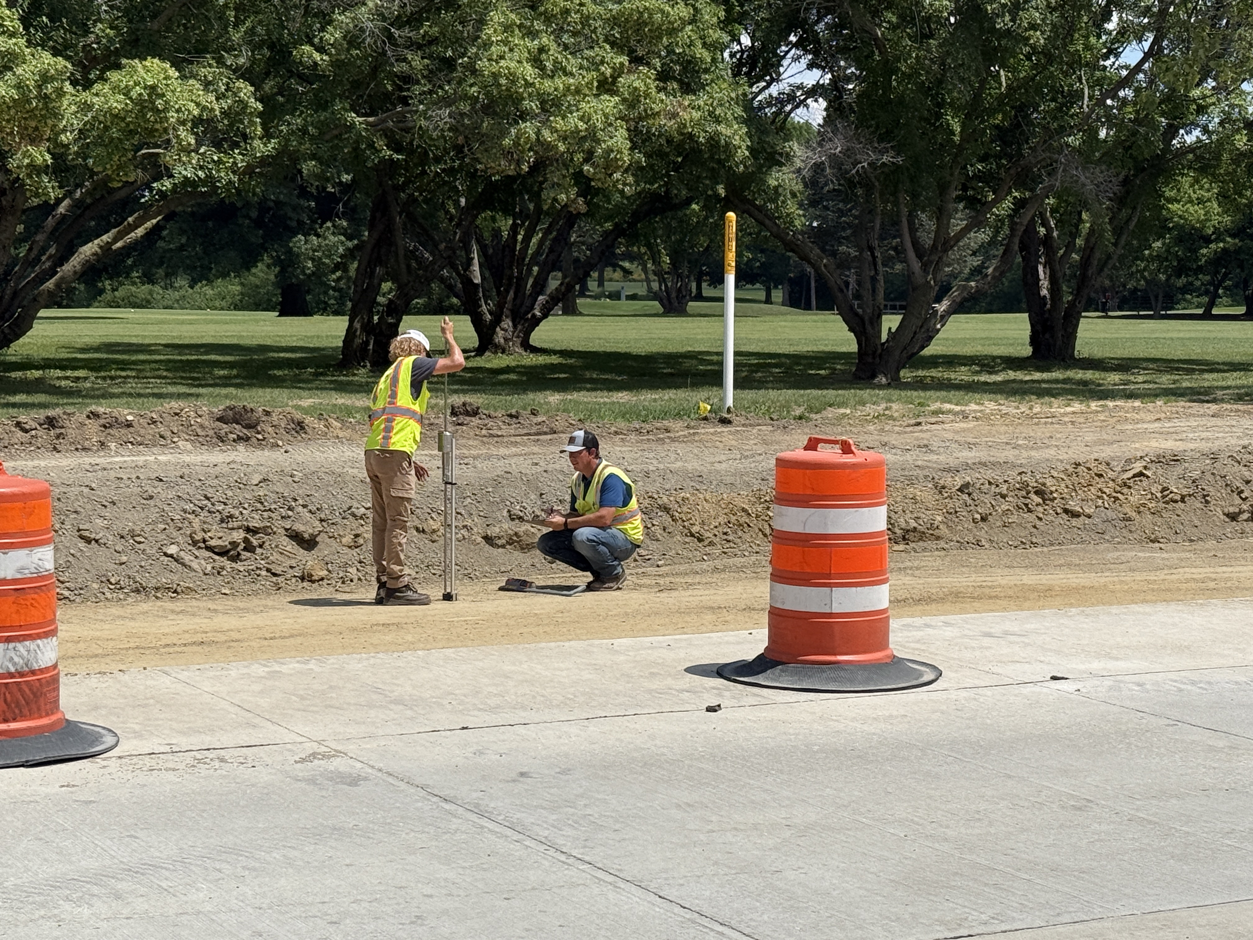 Two construction workers in safety vests working on a road construction site, with orange traffic barrels and trees in the background.