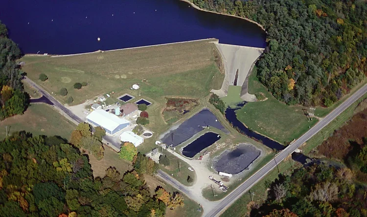 Aerial view of a dam with a water reservoir, hydroelectric power station, water intake structures, and a surrounding forested area.