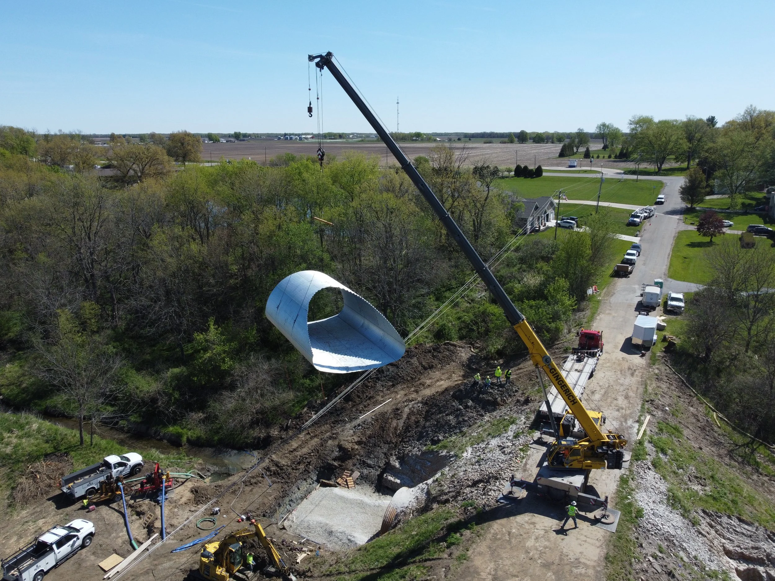 Road construction workers paving asphalt on a street in a residential neighborhood with houses, trees, utility poles, and orange traffic barrels.