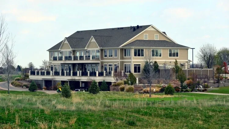 Large multi-story building with beige siding, multiple balconies, and a peaked roof, surrounded by a well-maintained lawn and landscaped yard.