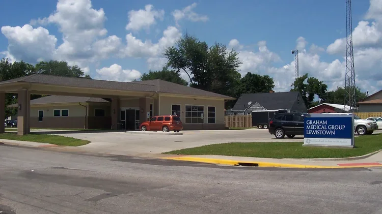 Exterior of Graham Medical Group Lewistown clinic with a parking lot, few cars, a blue sign, and a partly cloudy sky.