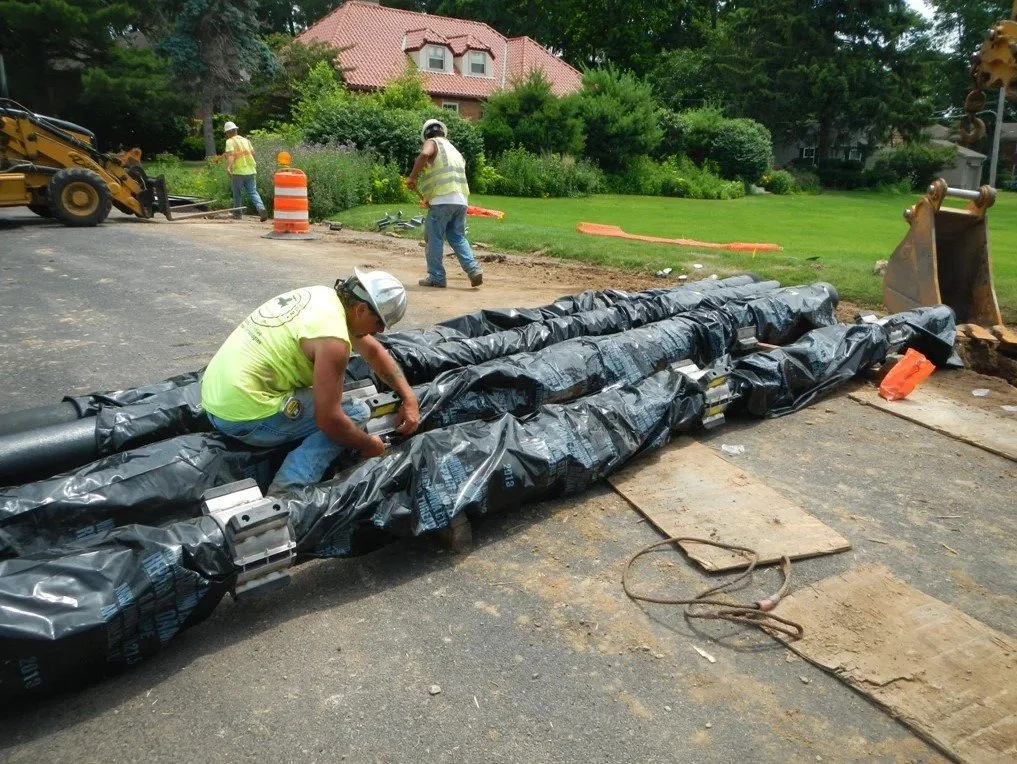 Construction workers installing a large pipeline on a street, with trees, houses, and construction equipment visible in the background.