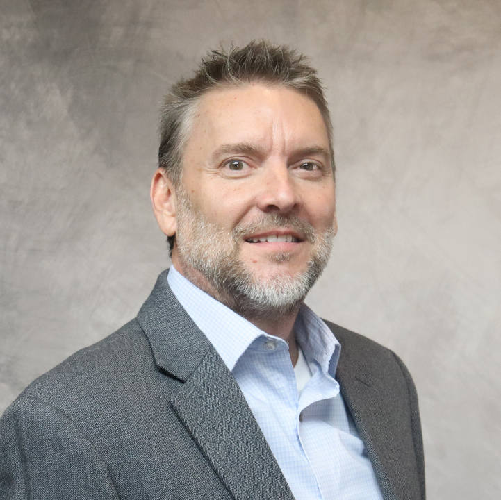 Portrait of a middle-aged man with gray hair and beard, wearing a gray suit jacket and light blue shirt, smiling at the camera against a neutral background.