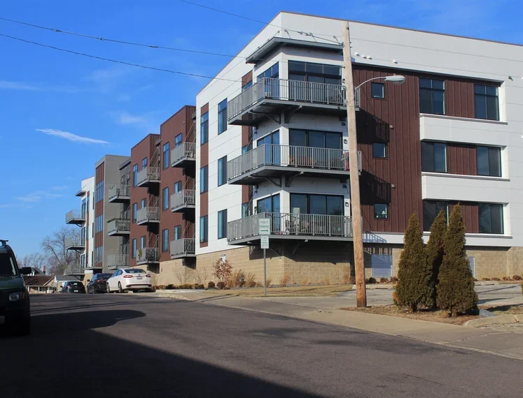 Multi-story apartment building with balconies, cars parked nearby, a utility pole, and some bushes in the foreground, under a blue sky.