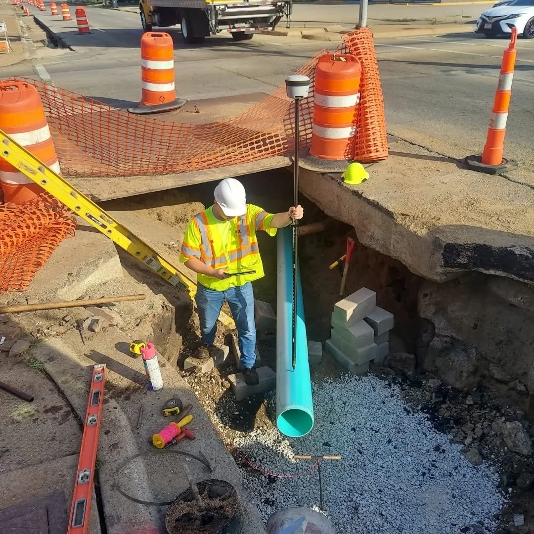 Construction worker in a yellow safety vest and white helmet installing a large light blue pipe in a deep excavation at a road construction site, surrounded by orange cones and orange safety barriers.