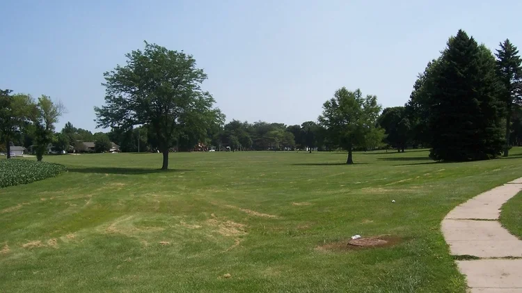 A park with a sidewalk, several trees, and open grassy areas under a clear blue sky.