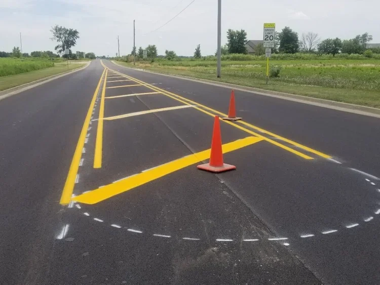 A newly paved road with traffic cones and fresh yellow road markings indicating a no-parking zone, with a speed limit sign of 20 miles per hour on the side of the road.