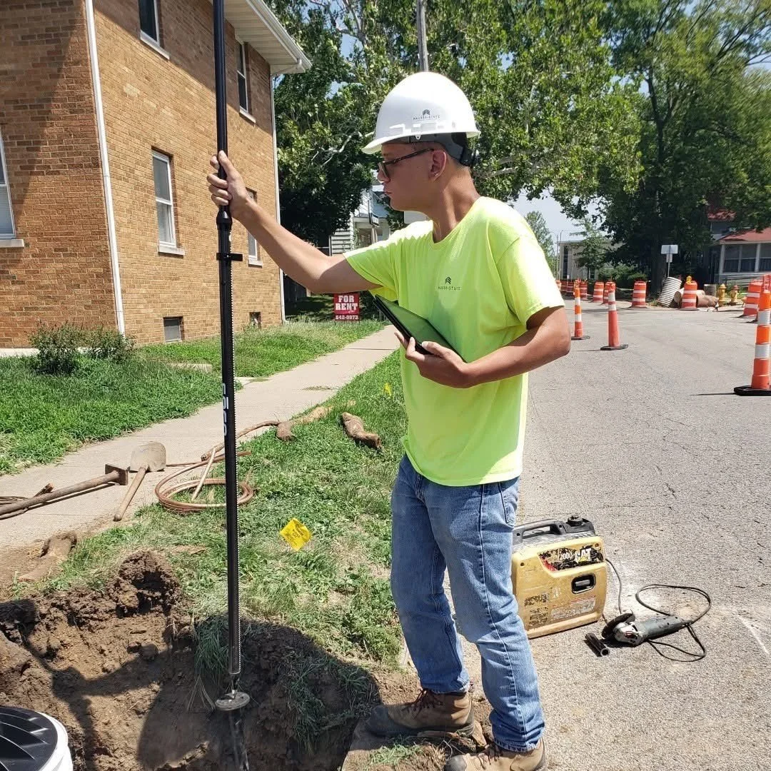 A construction worker wearing a white safety helmet, yellow safety shirt, and blue jeans operates a soil testing device near a sidewalk with a brick building behind, with orange traffic cones on the street and trees in the background.