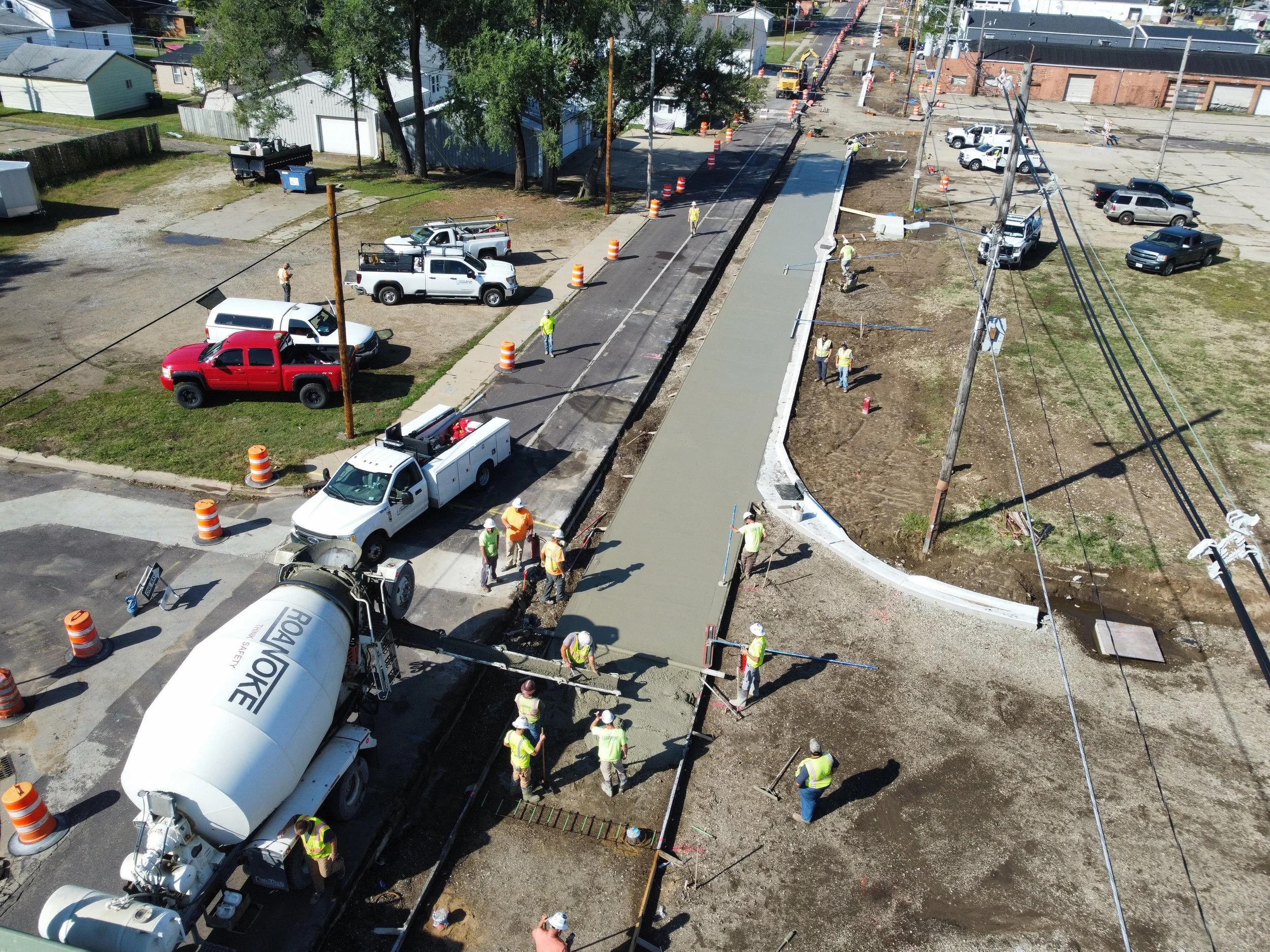 A road with orange traffic barrels indicating construction or maintenance work on the side of the road, yellow double center lines, overhead power lines, green trees, and a blue sky with some clouds.