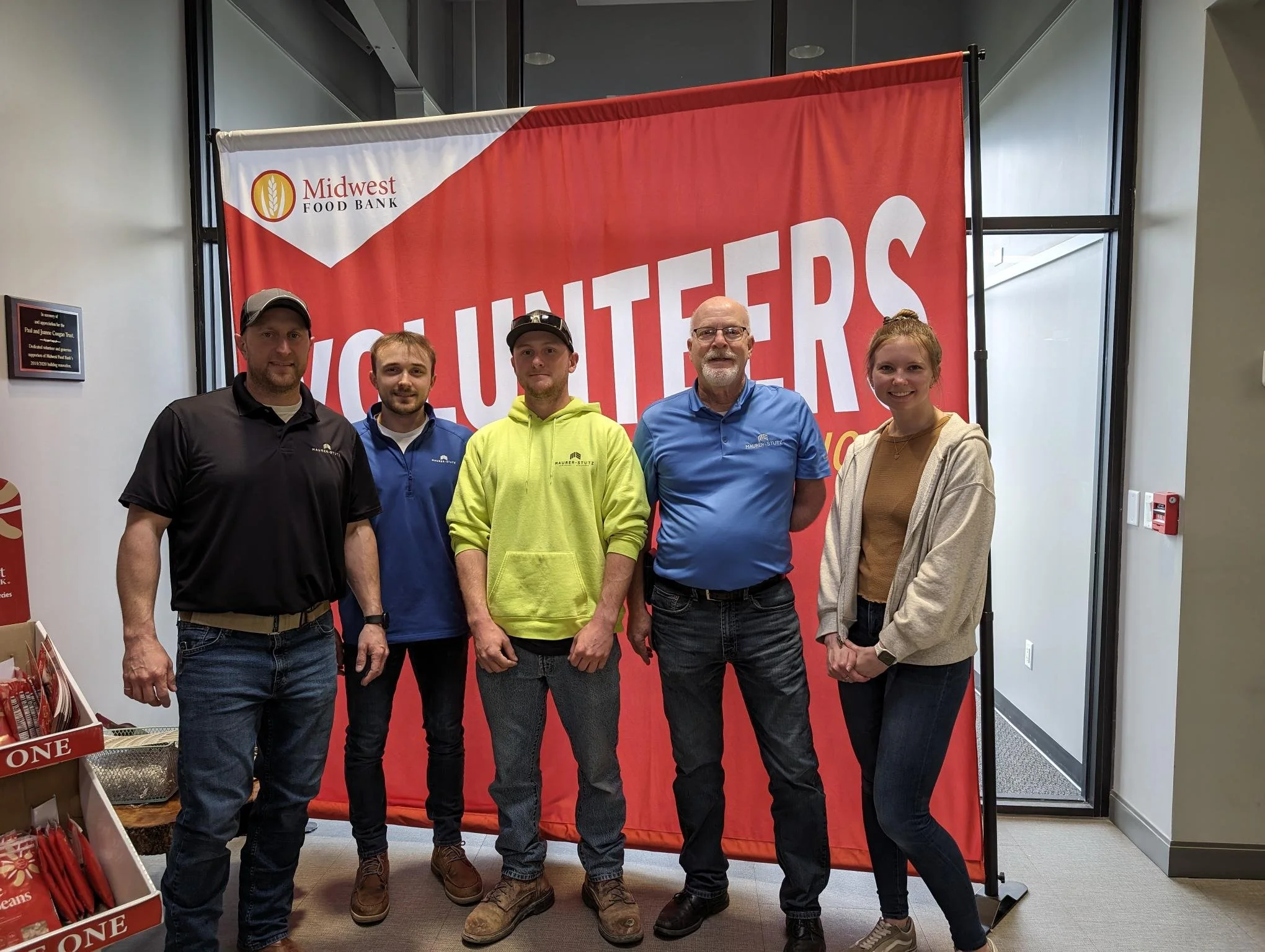 Group of five volunteers standing in front of a red Midwest Food Bank banner, indoors, smiling for the camera.