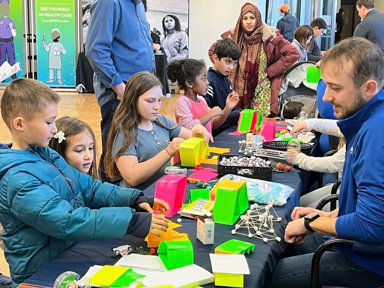 Children and adults participate in a science or craft activity at a table, with colorful materials like plastic shapes and models, in a room with informational posters and banners.