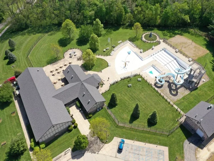 Aerial view of a large backyard featuring a water park with water slides, a pool, and lounging areas, surrounded by trees and a lawn