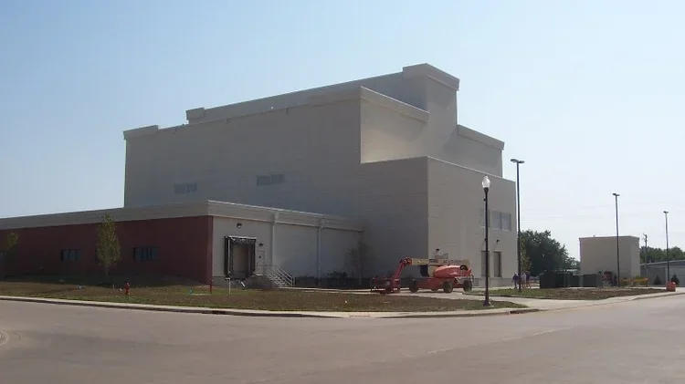 A large white industrial or warehouse building with a smaller red section in the front, on a paved street with lampposts and a few trees.