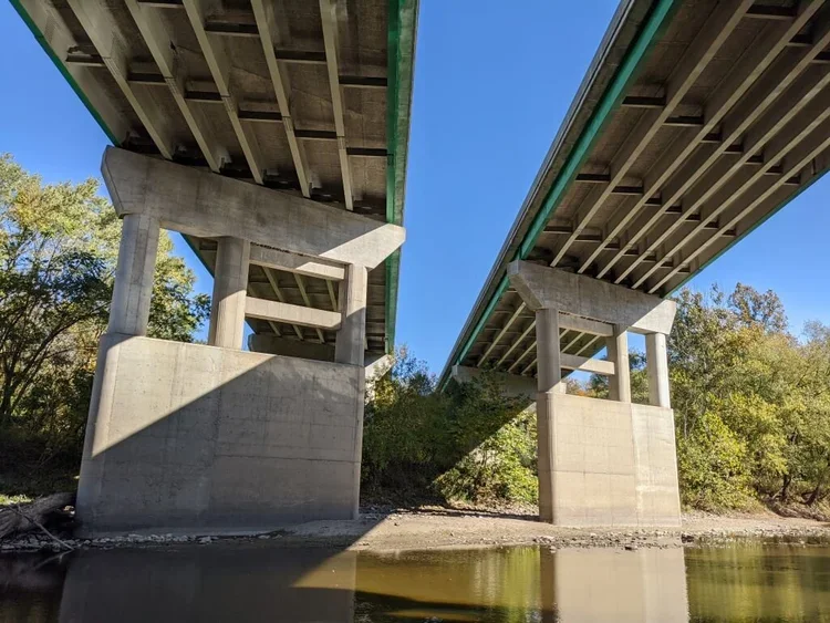 Looking up at two bridges supported by concrete pillars over a body of water, with trees in the background.