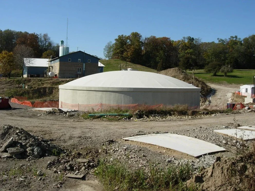 Construction site with a large white domed structure in the foreground, surrounded by dirt, construction materials, and orange safety barriers. Background features trees, a house, and a hill.