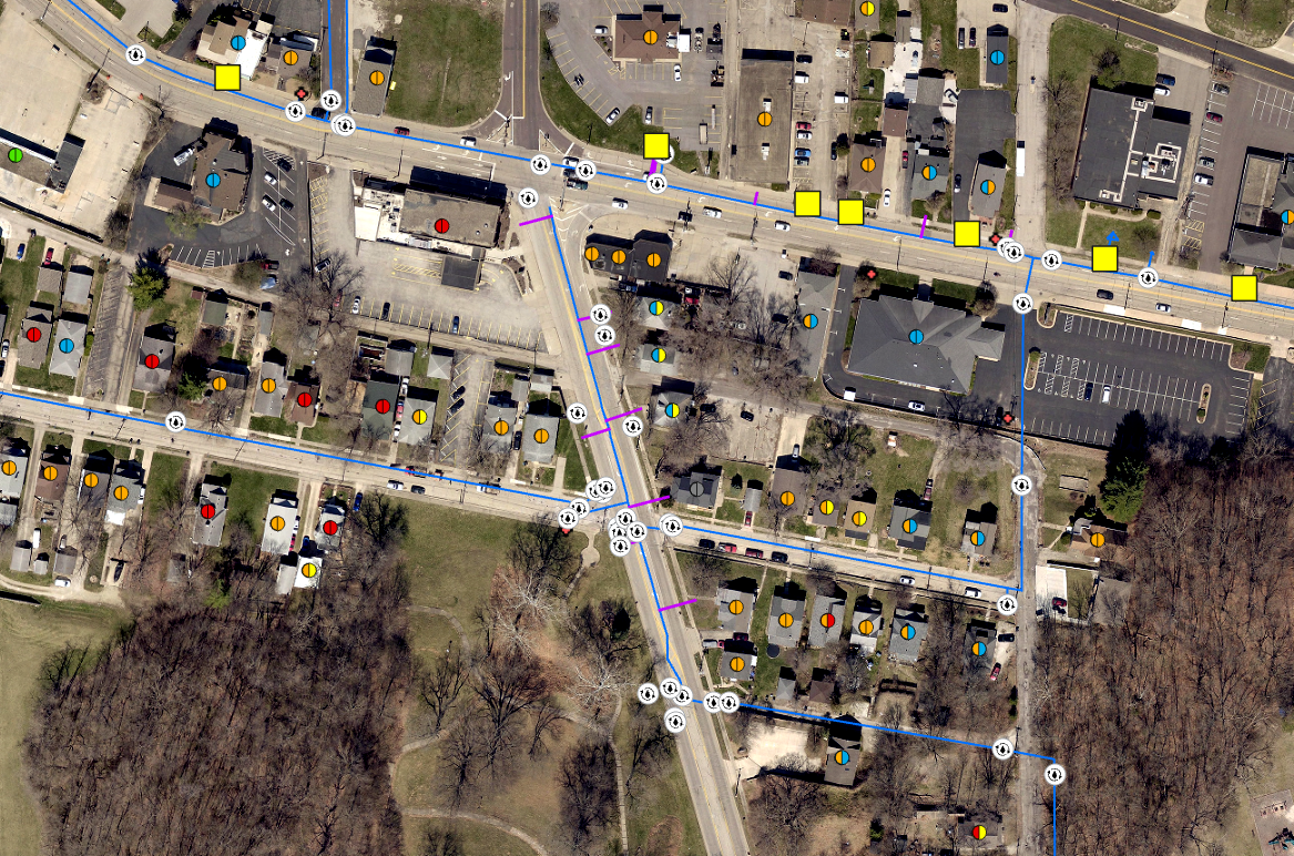  aerial view of a neighborhood with houses, a parking lot, and a street marked with blue lines and symbols indicating utilities or infrastructure.