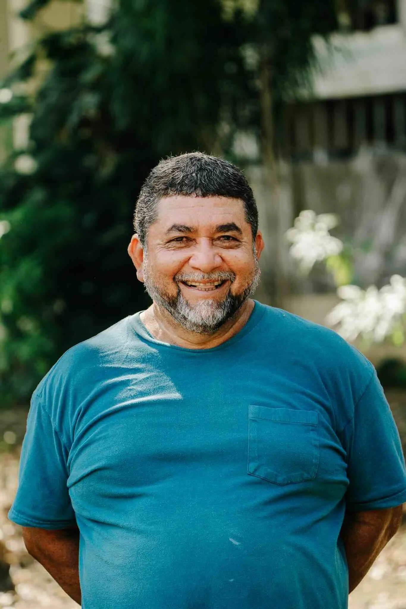 Smiling man with short dark hair and beard, wearing a teal t-shirt, standing outdoors with green foliage in the background.