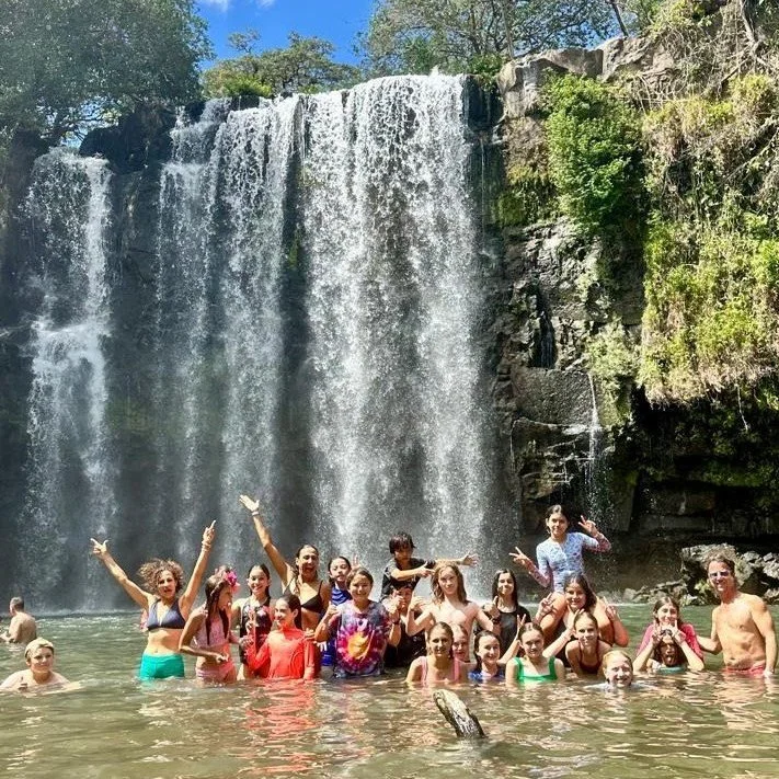 Group of high school students swimming and playing in a river at the base of a large waterfall on a sunny day during a field trip in Costa Rica