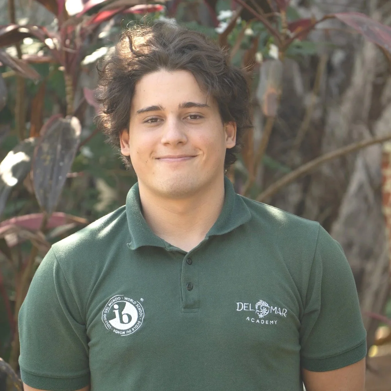 A young man with wavy brown hair, wearing a green polo shirt with logos, standing outdoors in front of plants with reddish leaves.