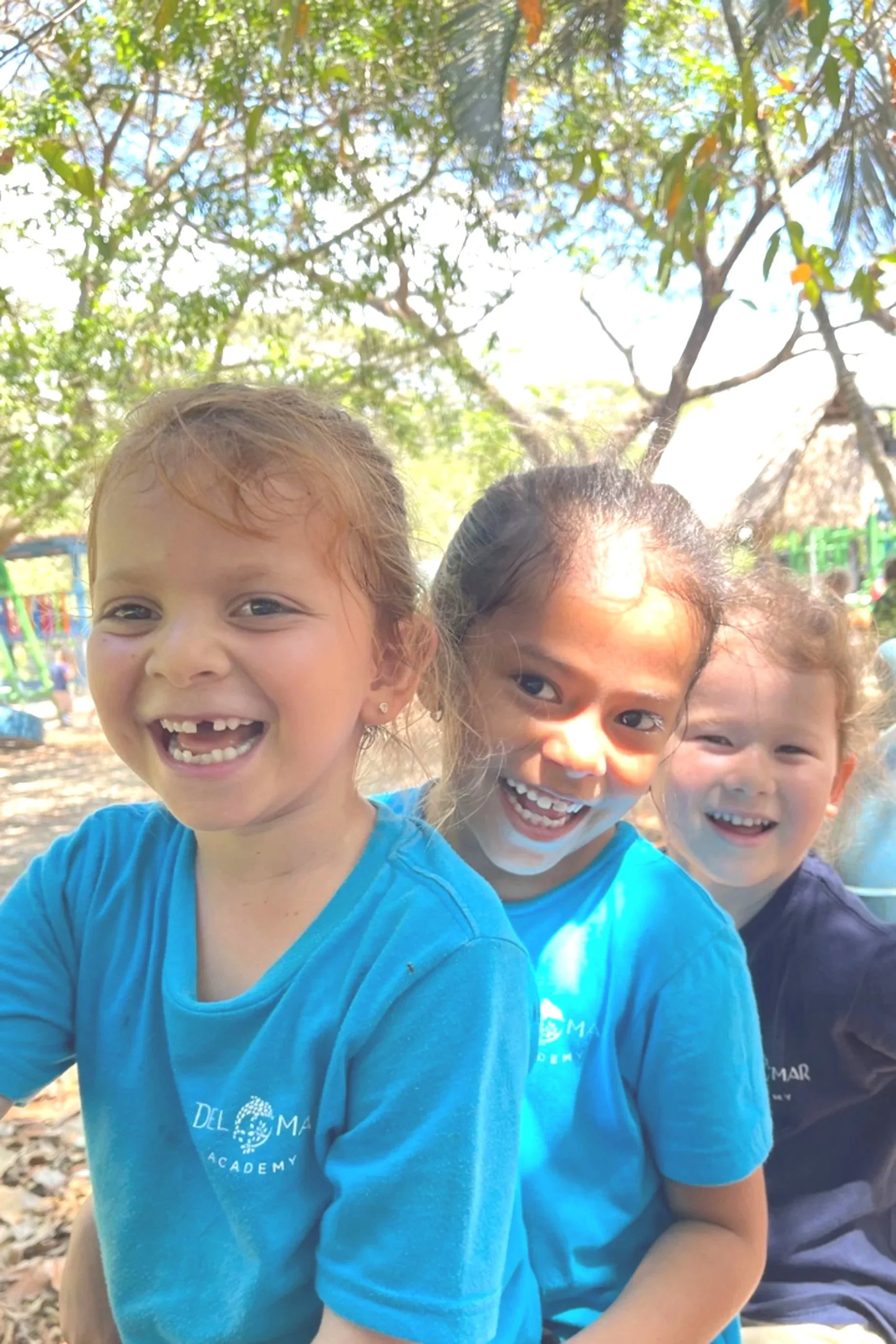 Three young girls smiling and happy outdoors with trees and a playground on Del Mar Academy's 11 acre school campus in Nosara Costa Rica
